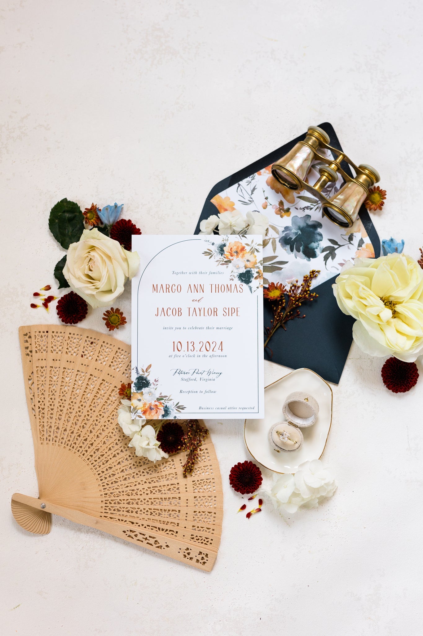 A flat lay of an invitation suite. The cards features a dark navy envelope with a floral liner and bold orange text on the main invite.
