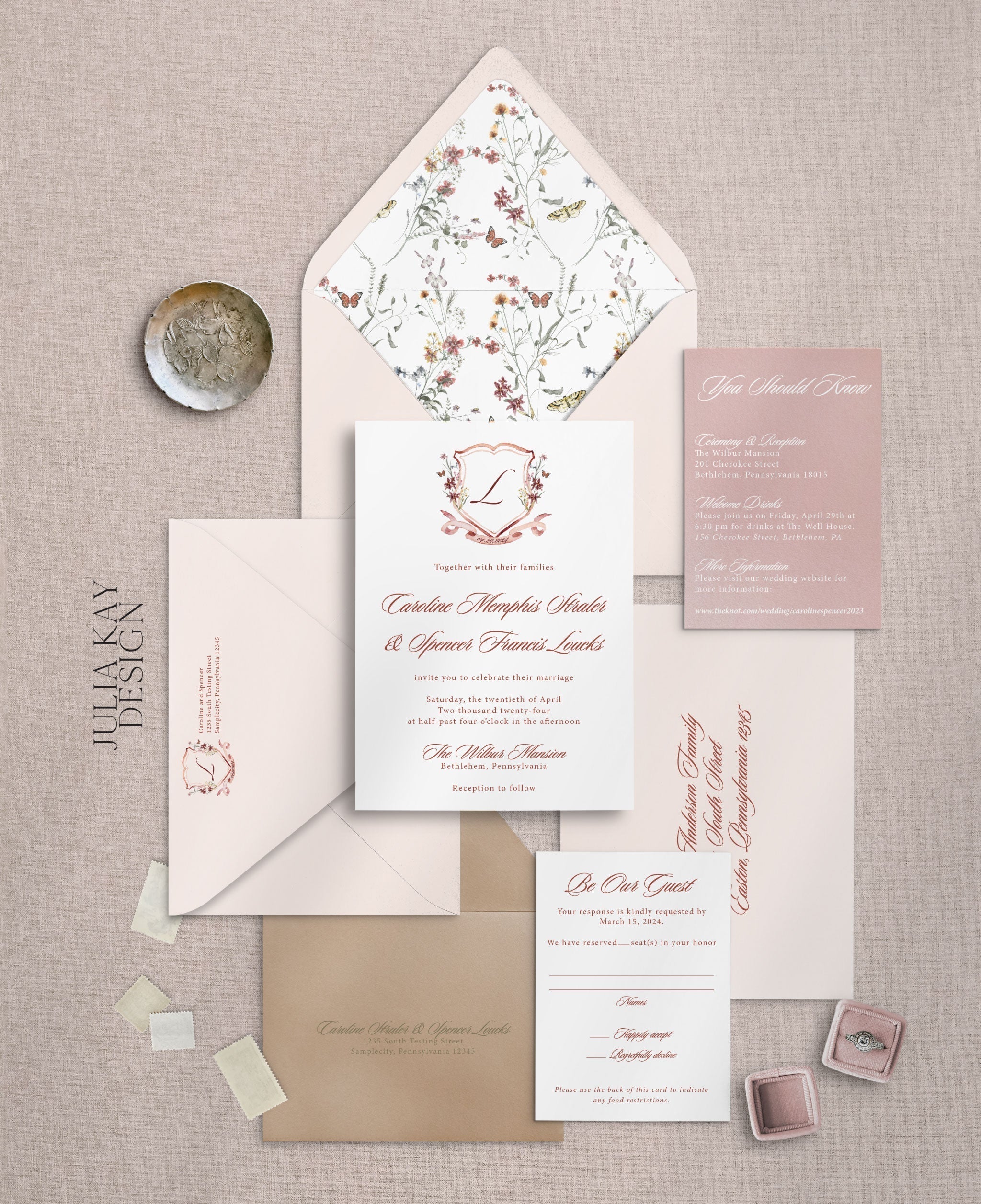 An invitation suite that has pink and rose colors. The center of the image has a white invitation card with red cursive writing. The top center features a crest with regal outlining and the letter "L" in the center.