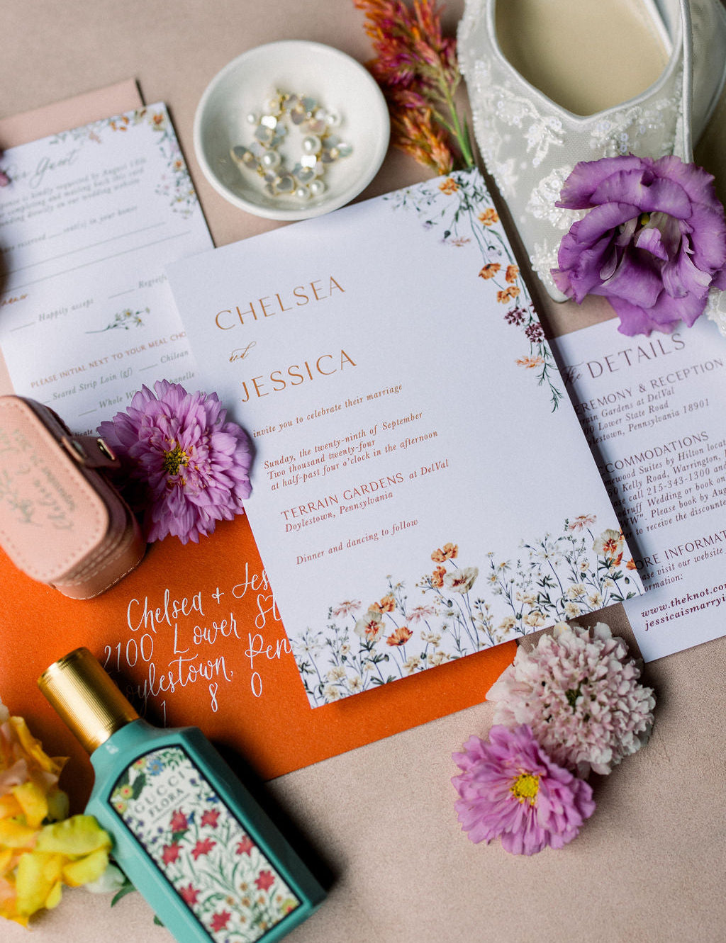 Flat lay of a wedding invitation suite.  The invite is on white card stock with orange print.  The card also features florals that run along the bottom and top right corner.  