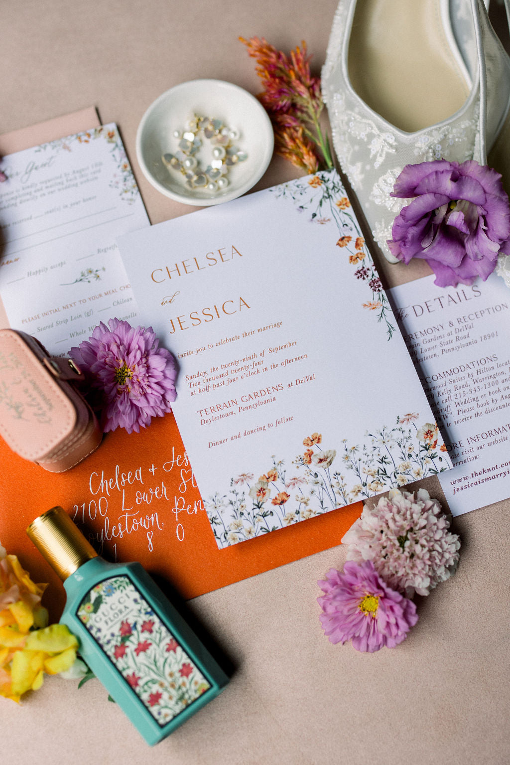 Flat lay of a wedding invitation suite.  The invite is on white card stock with orange print.  The card also features florals that run along the bottom and top right corner.  