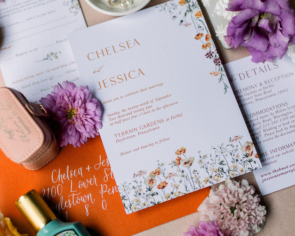 Flat lay of a wedding invitation suite.  The invite is on white card stock with orange print.  The card also features florals that run along the bottom and top right corner.  