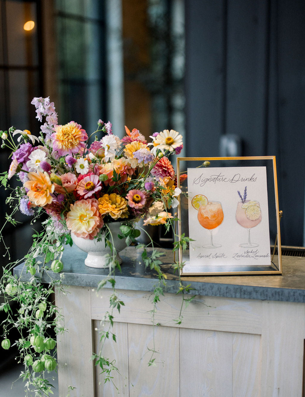 A white sign with illustrations of cocktail drinks being served at a wedding bar.  The poster is placed in a gold frame and is displayed next to a floral arrangement. 