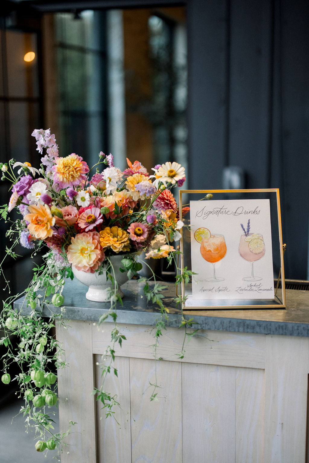 A white sign with illustrations of cocktail drinks being served at a wedding bar.  The poster is placed in a gold frame and is displayed next to a floral arrangement. 
