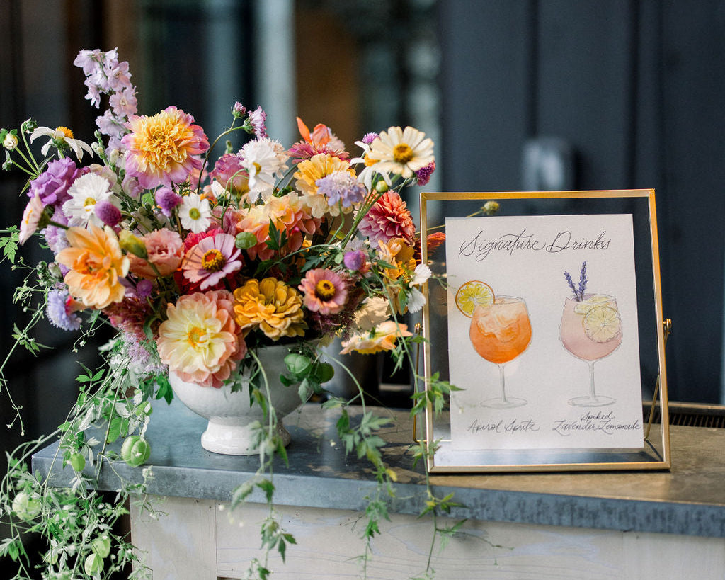 A white sign with illustrations of cocktail drinks being served at a wedding bar.  The poster is placed in a gold frame and is displayed next to a floral arrangement. 
