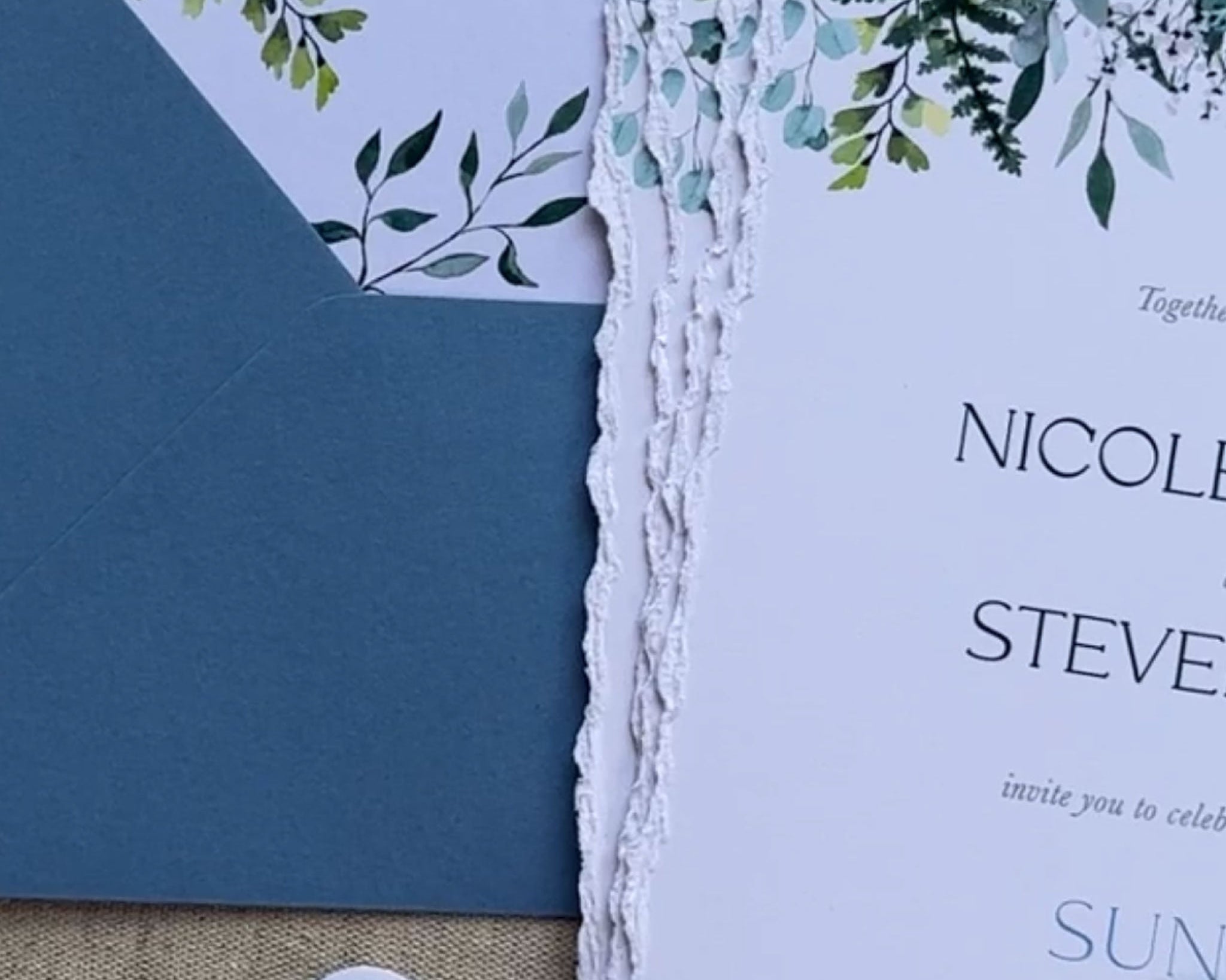 An invitation suite with greenery on the top of the suite.  The edges of the invites have a deckled edge.