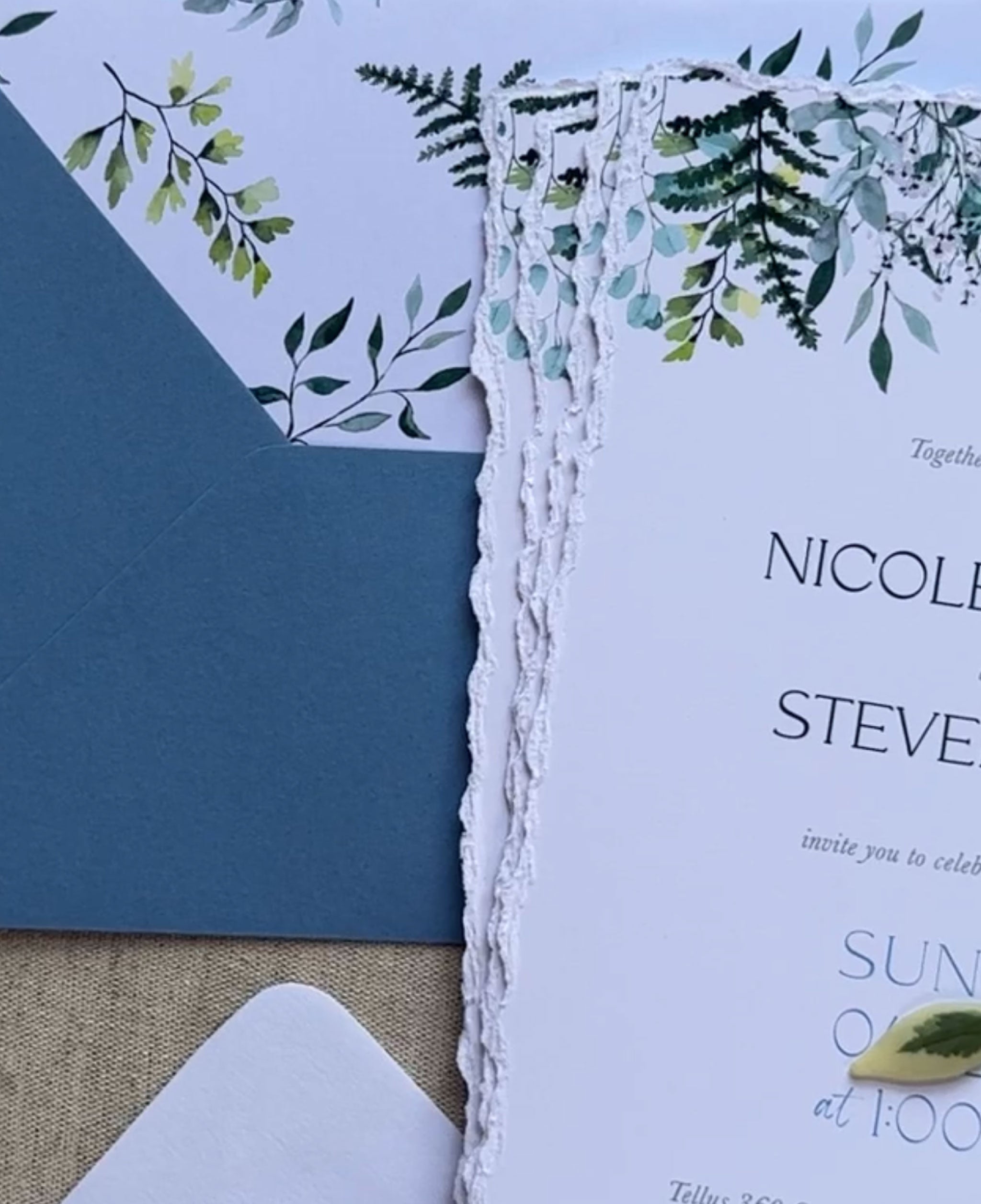An invitation suite with greenery on the top of the suite.  The edges of the invites have a deckled edge.