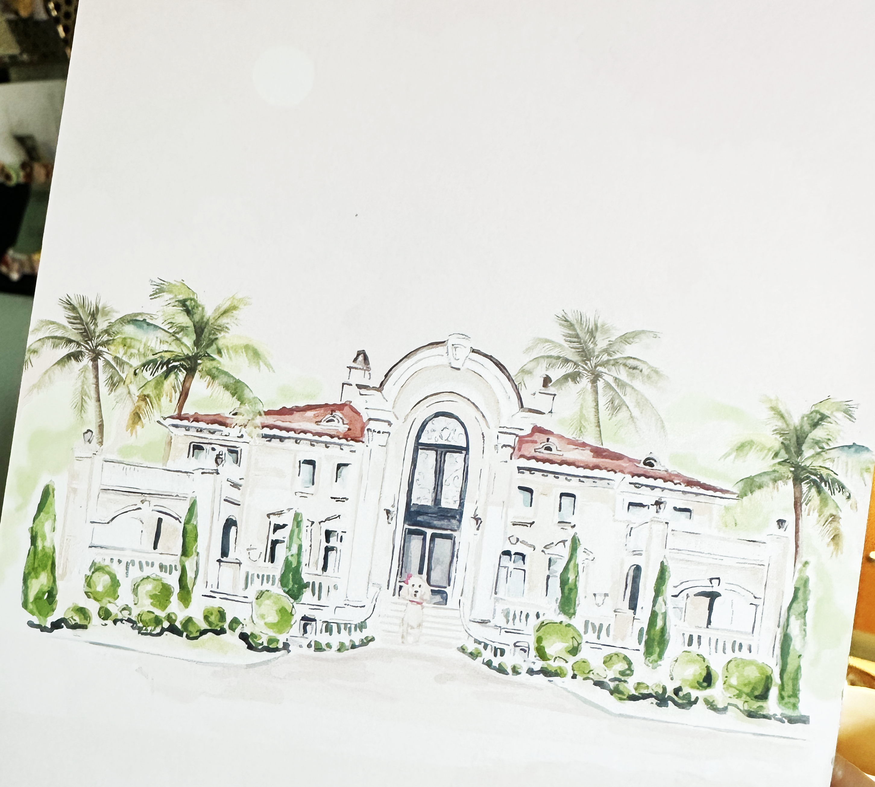 Water color illustration of a Spanish styled mansion.  The venue has red tile roofing and on the front steps a white poodle with a red bow is sitting.