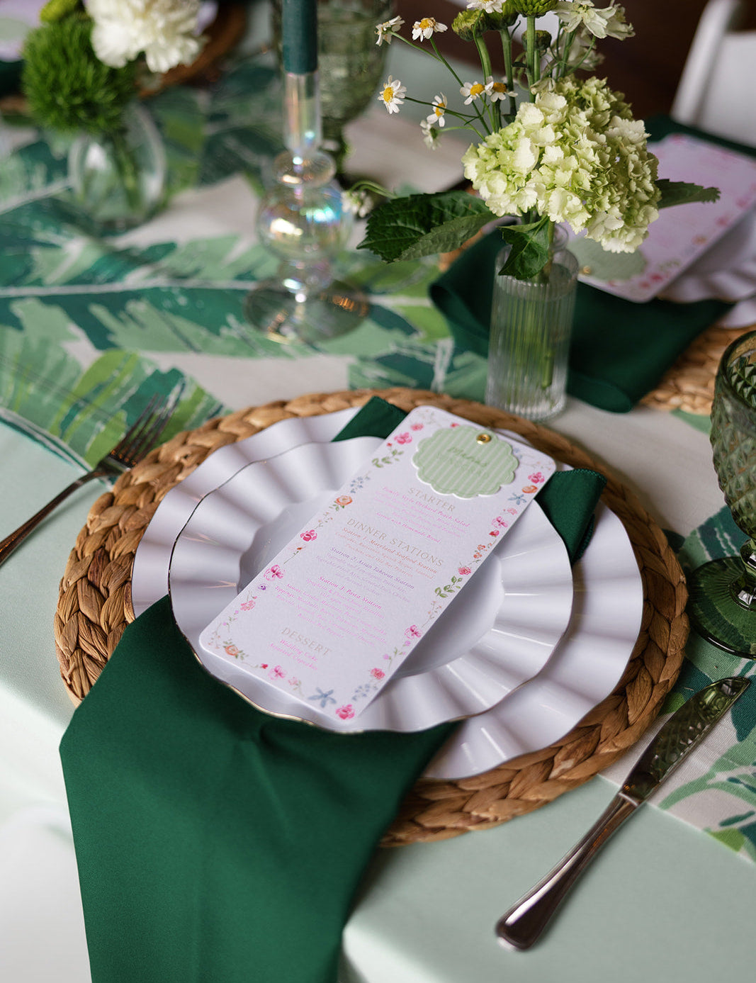 A tablescape with green florals, linens, and napkins. On stop of stacked plates is a dinner menu.