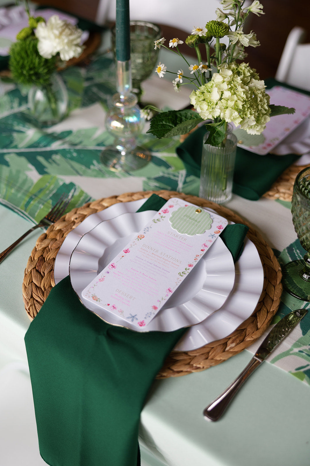 A tablescape with green florals, linens, and napkins. On stop of stacked plates is a dinner menu.