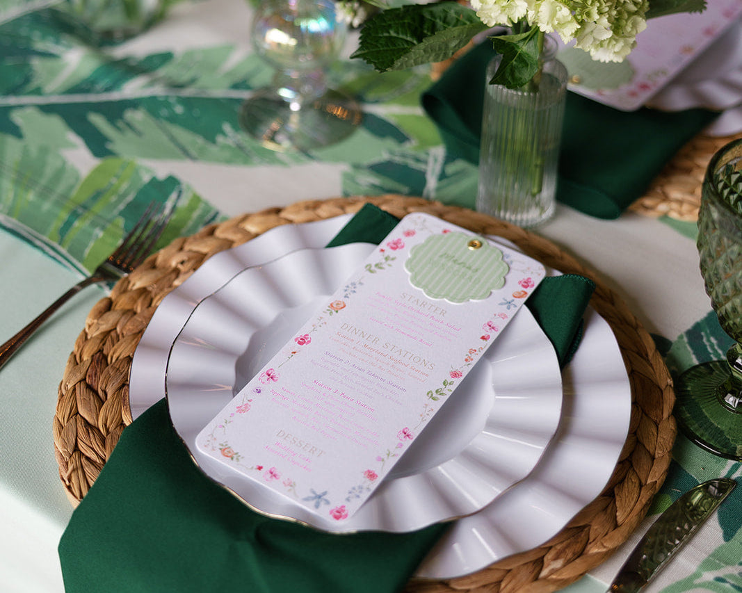 A tablescape with green florals, linens, and napkins. On stop of stacked plates is a dinner menu.
