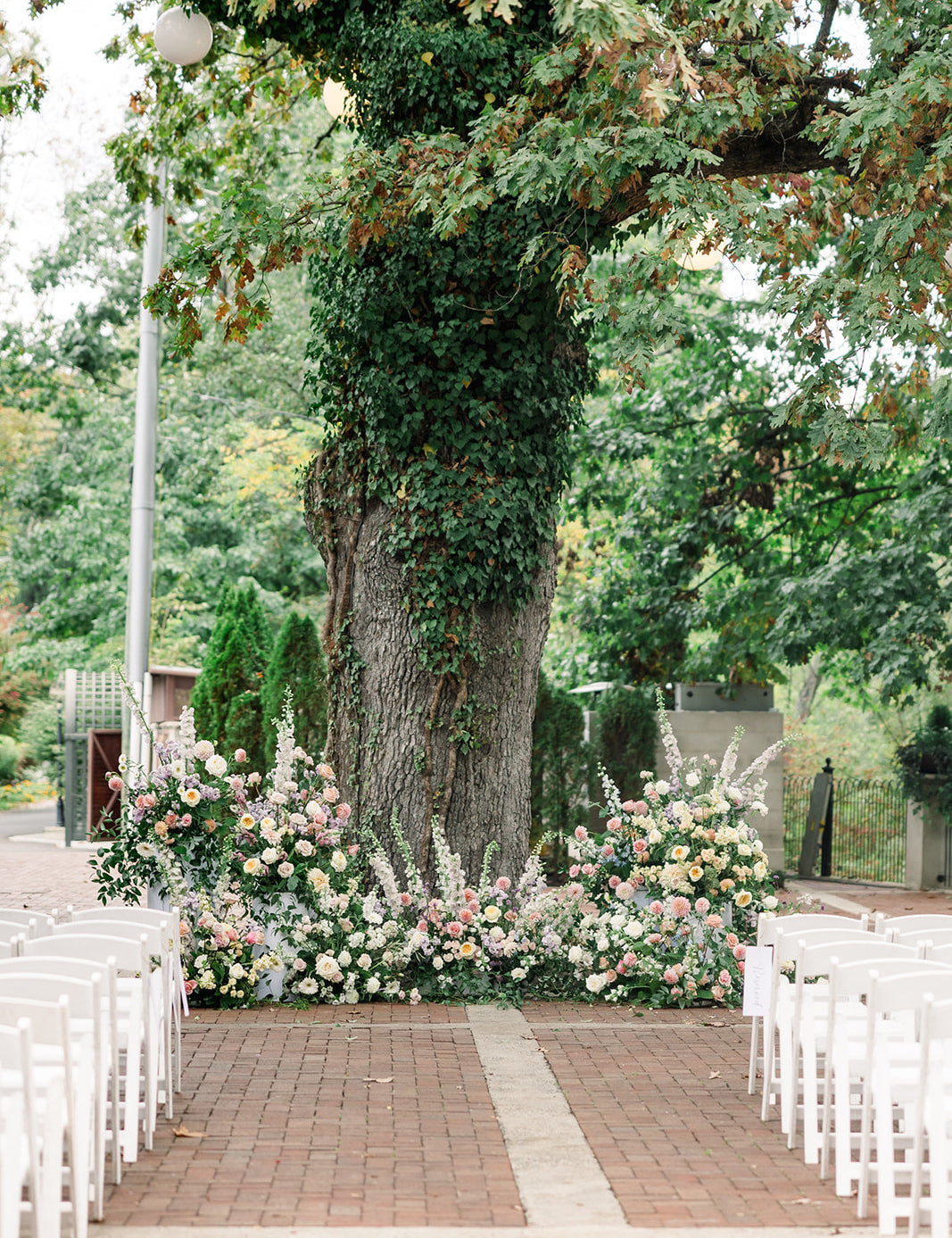 Image of an outdoor wedding venue.  The image looks down a brick isle to a large oak tree.  The tree is with wrapped in vines and pastel florals are placed at the base of the tree. Rows of white chairs sit on both sides of the isle.