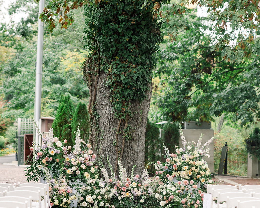 Image of an outdoor wedding venue.  The image looks down a brick isle to a large oak tree.  The tree is with wrapped in vines and pastel florals are placed at the base of the tree. Rows of white chairs sit on both sides of the isle.