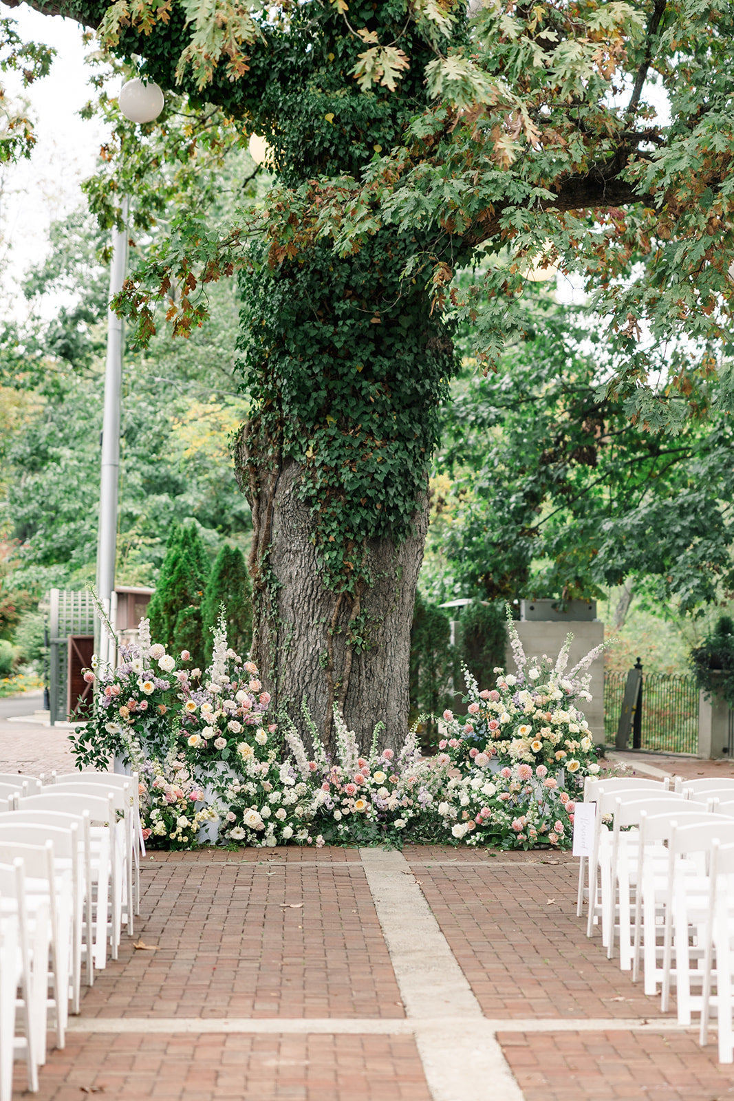 Image of an outdoor wedding venue.  The image looks down a brick isle to a large oak tree.  The tree is with wrapped in vines and pastel florals are placed at the base of the tree. Rows of white chairs sit on both sides of the isle.
