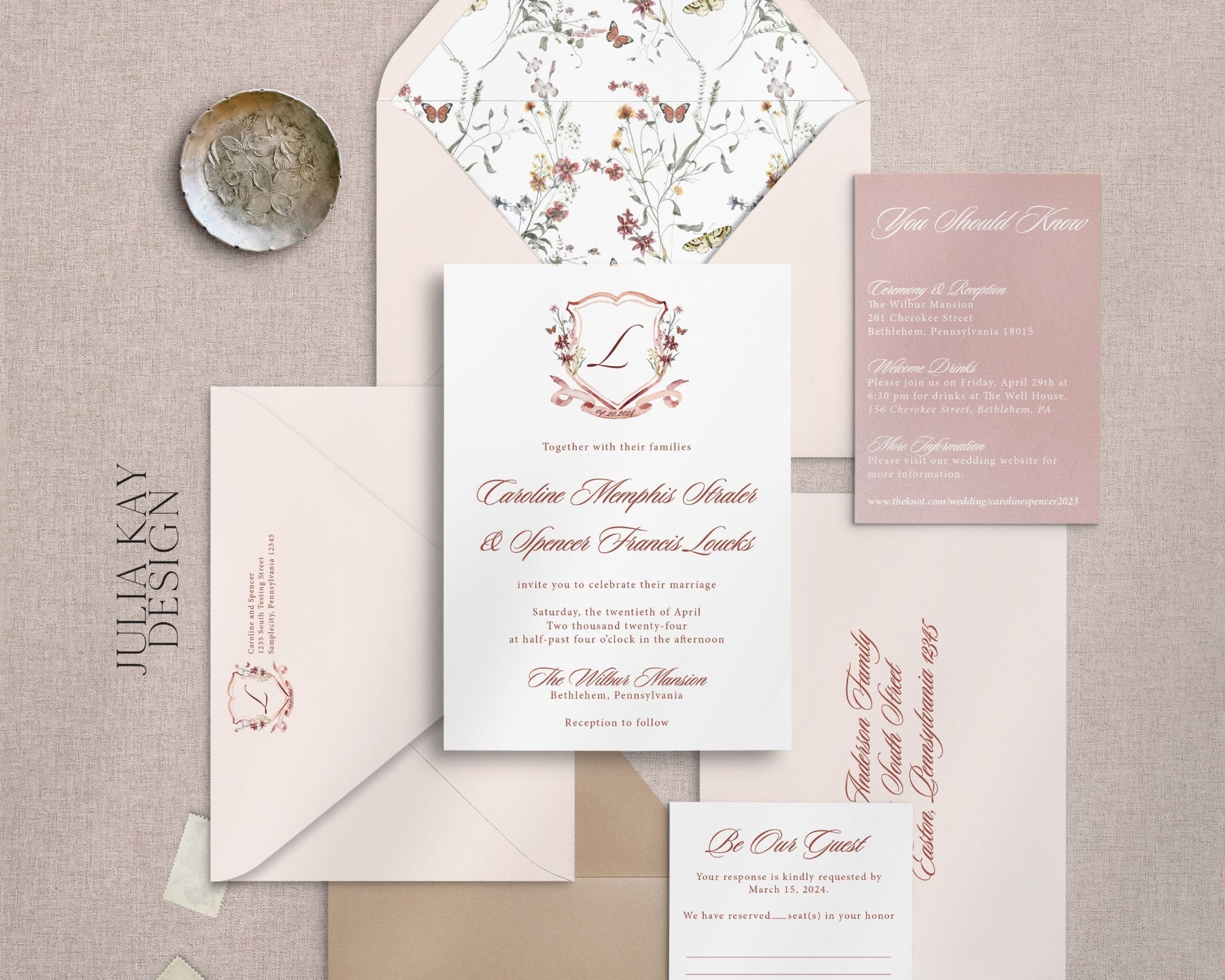 An invitation suite that has pink and rose colors.  The center of the image has a white invitation card with red cursive writing.  The top center features a crest with regal outlining and the letter "L" in the center. 