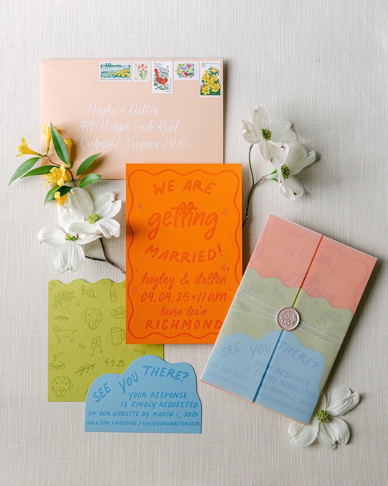 A styled flat-lay of a handmade paper invitation suite with pastel peach, bright orange, sage green, and sky blue cards, flowers, and a wax seal on a light wood background.