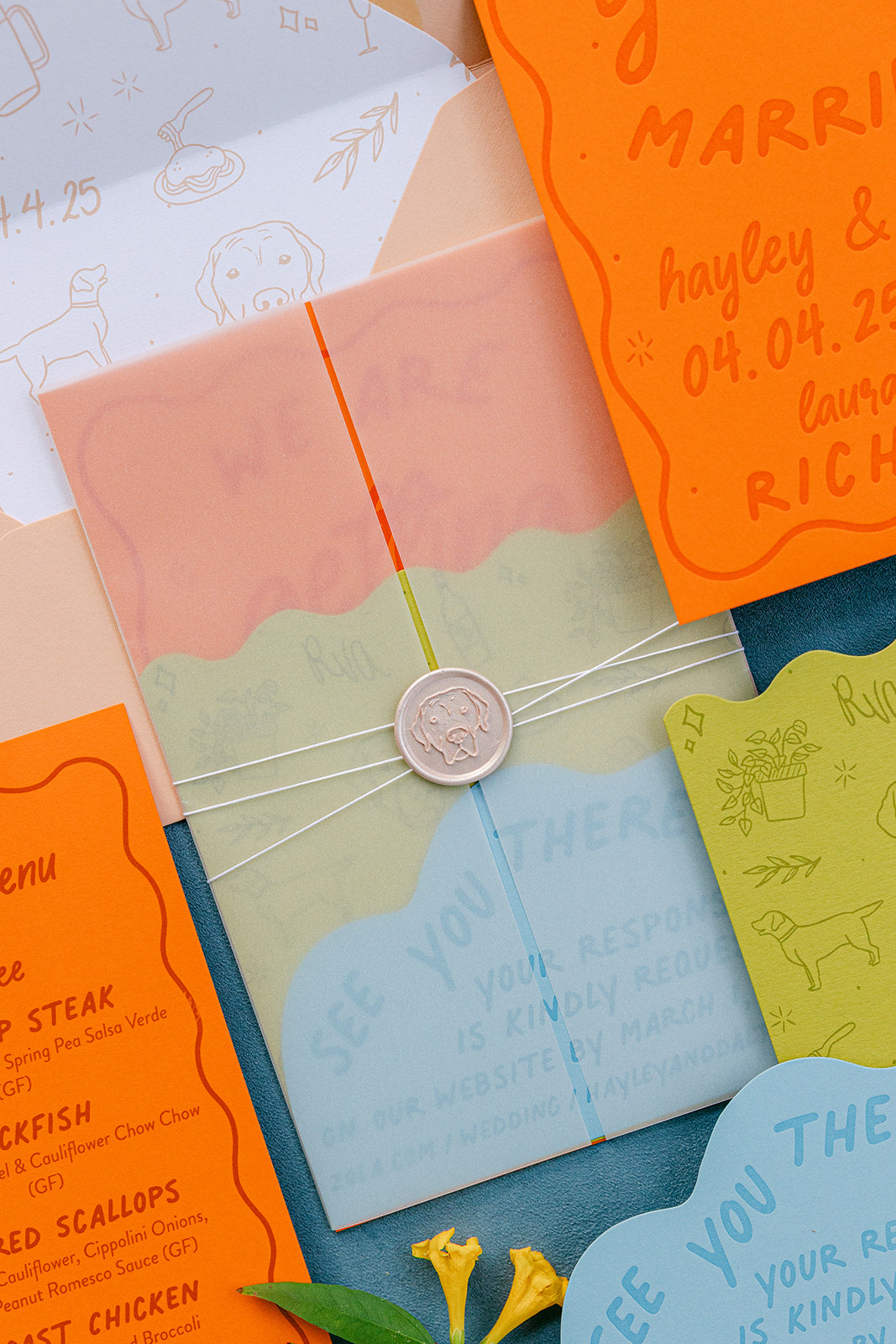 A styled flat-lay of a colorful wedding invitation suite featuring pastel and bright orange envelopes, cards, and a wax seal on a textured blue surface.