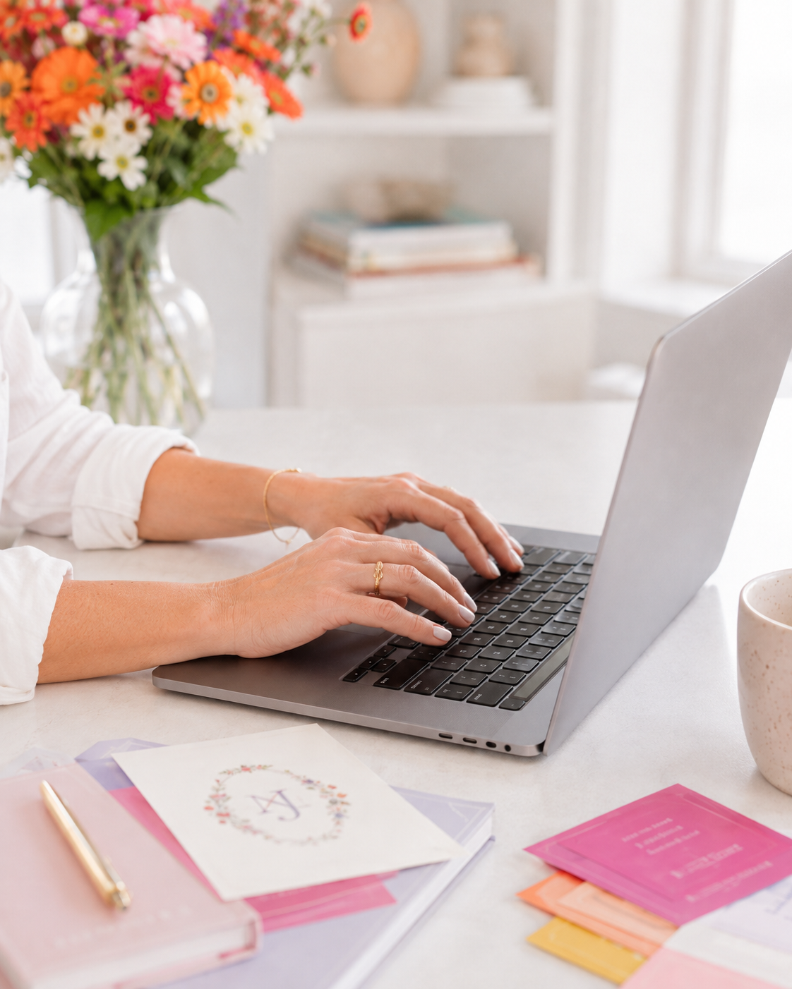 A person typing on a silver laptop at a white desk, with colorful stationery, a vase of flowers, and a mug in a bright, softly blurred home office setting.