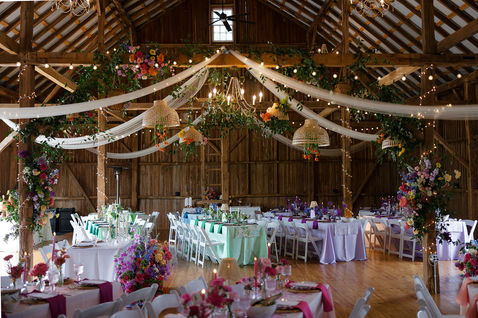 A rustic barn event space decorated for a wedding or reception, with round tables set in white, lavender, and mint green tablecloths, surrounded by white chairs. The venue features a high wooden beam ceiling adorned with draped white fabric, lush green garlands, colorful floral arrangements, and warm string lights.