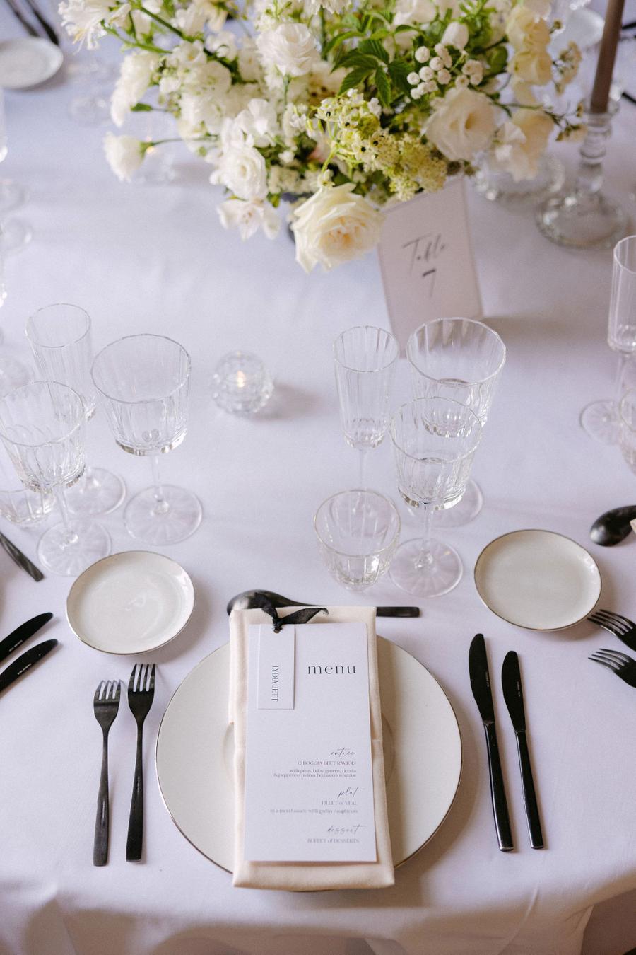 A styled wedding table setting with white plates, black cutlery, clear glassware, and a cream linen napkin with a printed menu, set on a white tablecloth with a floral centerpiece in the background.
