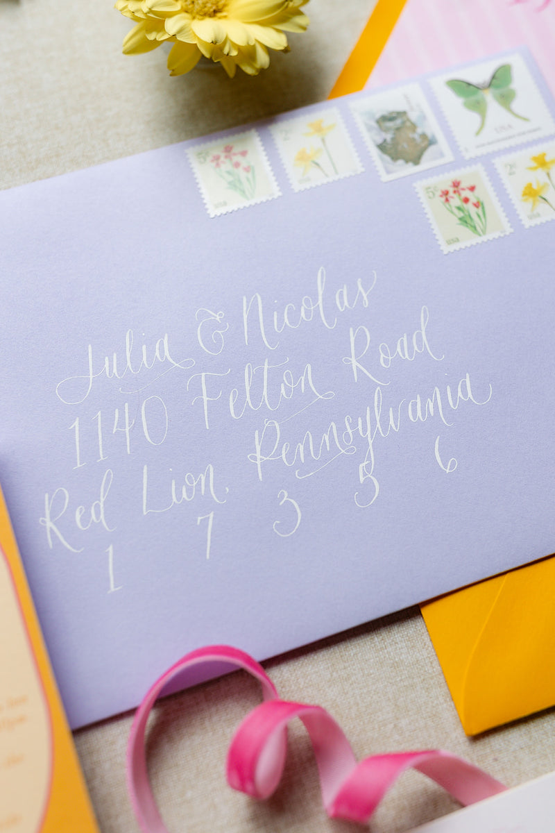 A lavender invitation envelope with elegant white script address writing and decorative postage stamp illustrations along the top edge, styled on a neutral fabric surface with a yellow flower, pink ribbon, and orange envelope.
