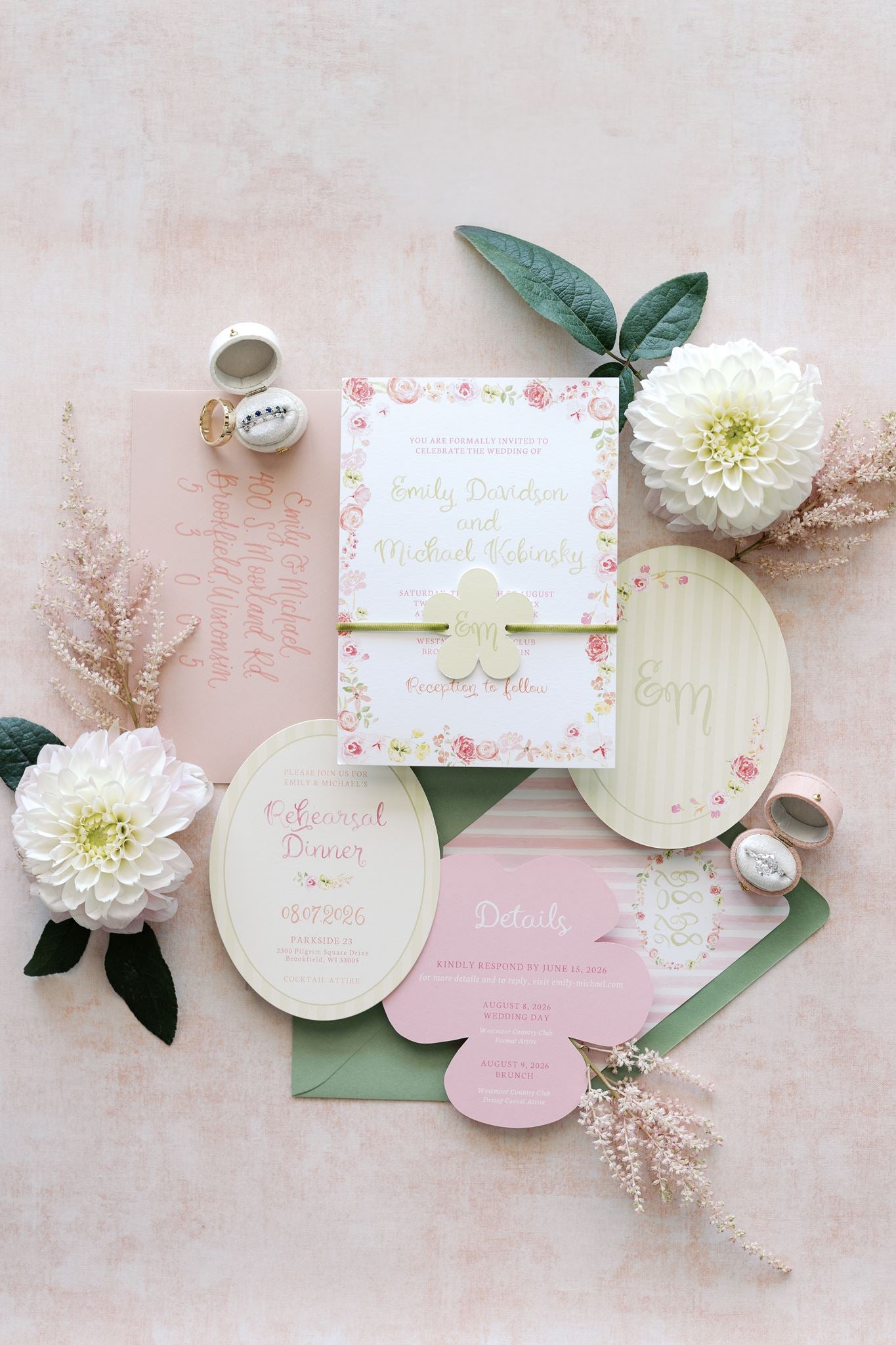 A styled flat lay of a wedding invitation suite on a soft blush pink background, featuring a floral square invitation, round and oval cards, green envelopes, dried flowers, and small metallic rings.