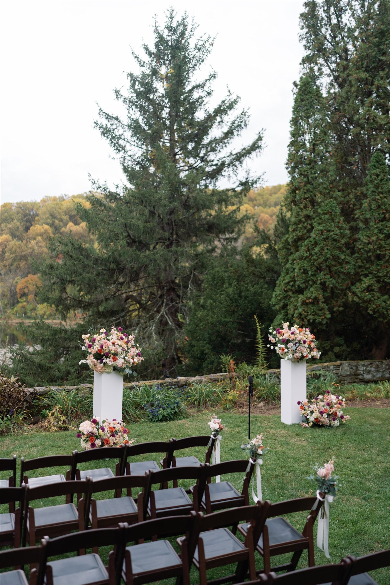 Outdoor wedding setup with floral arrangements on white pillars and chairs in a forested area.