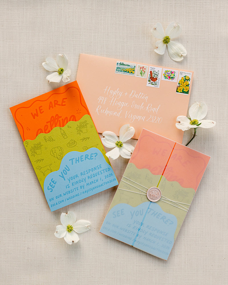 A styled flat-lay of a rustic wedding invitation suite on a textured linen background, surrounded by white dogwood blossoms. The suite includes a peach-colored envelope addressed to 'Hagley + Dalton', a bright orange and lime-green 'We Are Getting' card, a blue 'See You There?' RSVP card, and a peach, sage, and light blue envelope tied with twine and a wax seal.