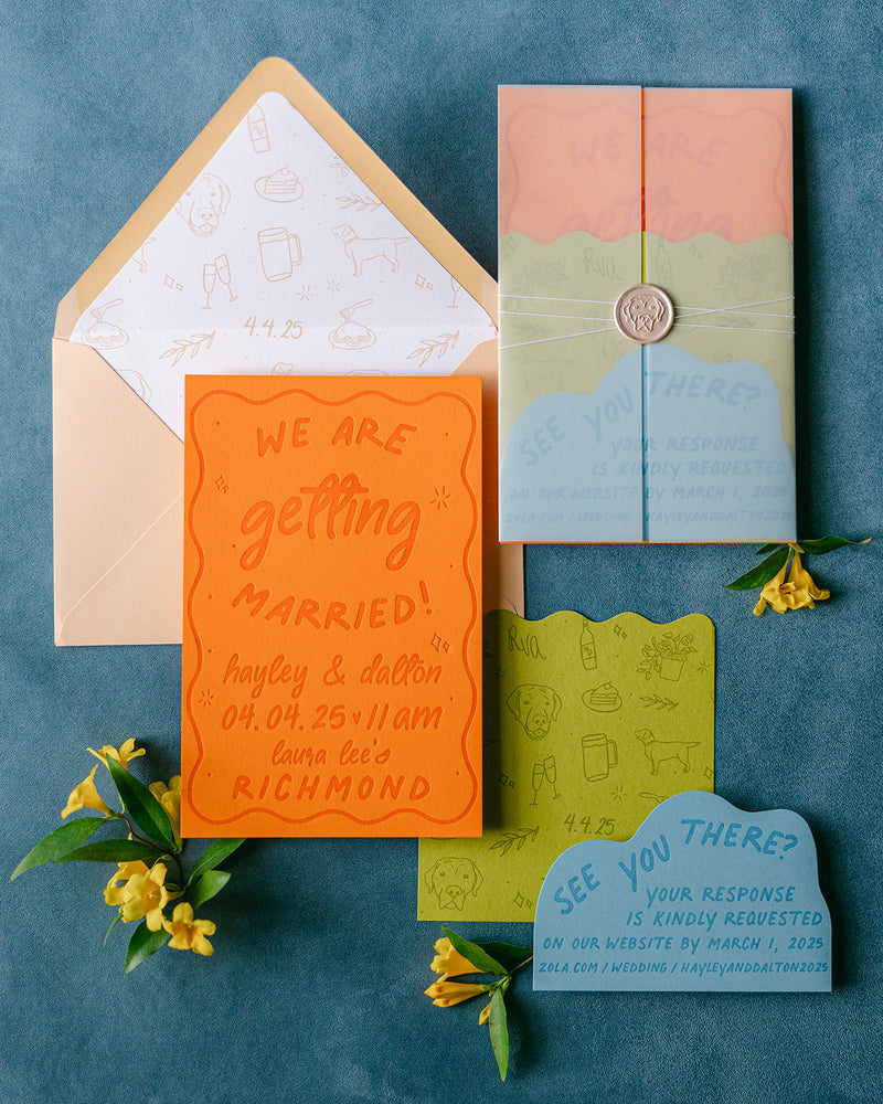 A coordinated wedding invitation suite arranged on a textured blue surface, including an orange 'we are getting married' card, a peach and blue 'See You There?' card tied with string and sealed with wax, a green scalloped card, a light blue 'See You There?' card, and a cream envelope with printed liner paper. Small yellow flowers and green leaves are placed around the items.