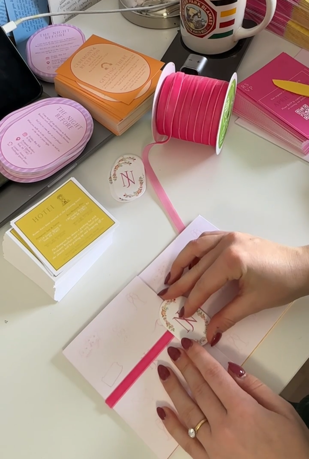 A close-up of hands applying a round decorative sticker to a pink ribbon notebook band, surrounded by stationery supplies like a spool of pink ribbon, business cards, and a mug on a white desk.