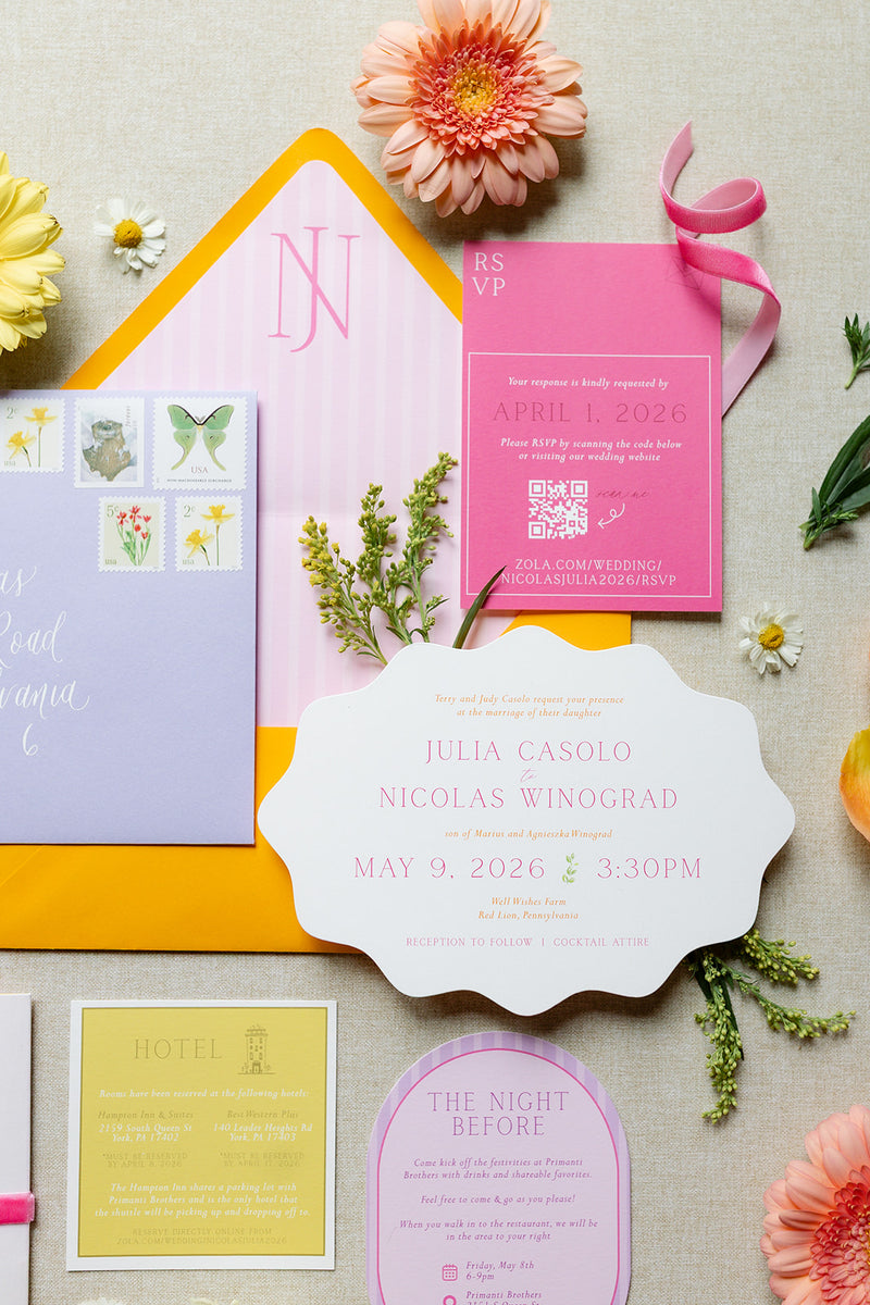 A styled flat-lay of a floral stationery suite on a light beige fabric background, featuring a pastel pink and lavender wedding invitation card, matching envelopes, a pink RSVP card, a yellow hotel card, and decorative flowers and greenery.