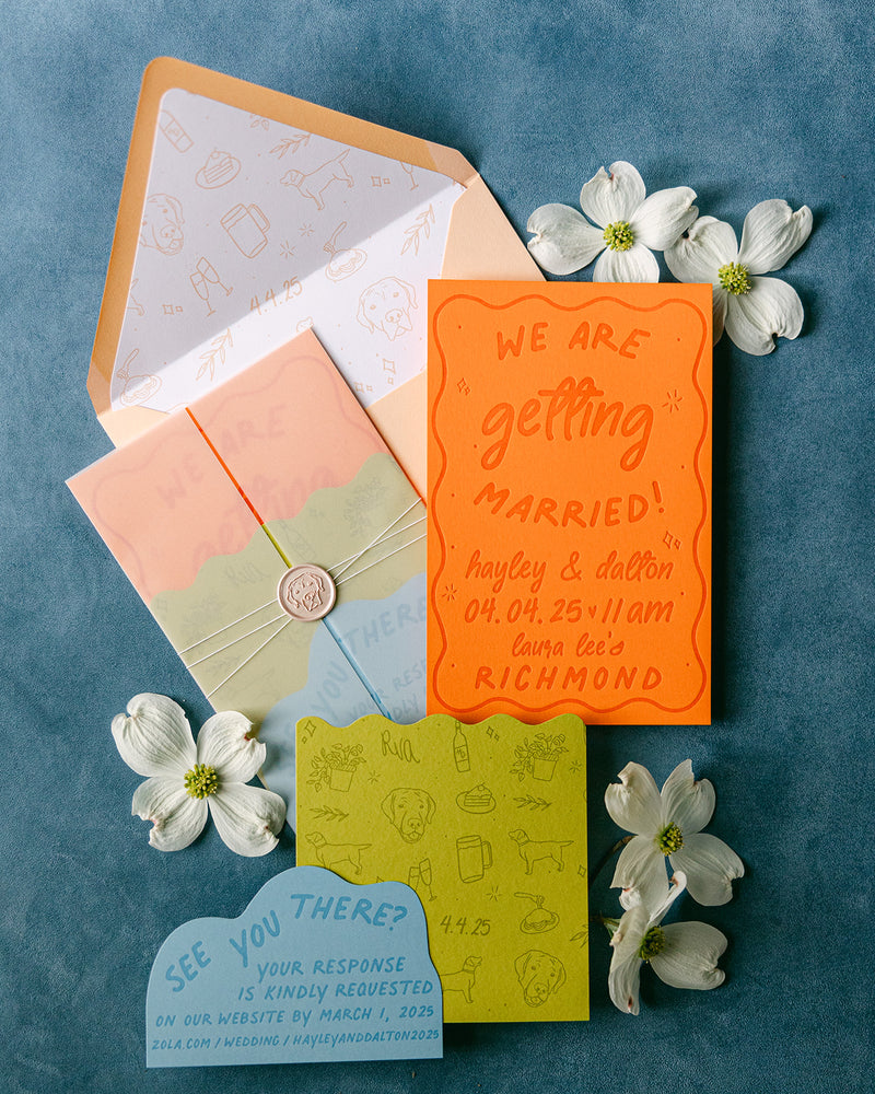 A styled flat-lay of a wedding invitation suite on a textured blue background, featuring an orange 'getting married' card, a peach and mint envelope, a pastel layered card tied with string and wax seal, a lime green RSVP card, and a blue 'see you there?' card, decorated with white dogwood flowers.