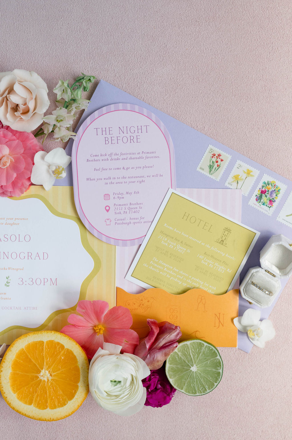A styled flat-lay of a wedding invitation suite on a dusty pink background, featuring a lavender envelope, a pink 'The Night Before' card, a yellow hotel card, floral accents, and citrus slices.