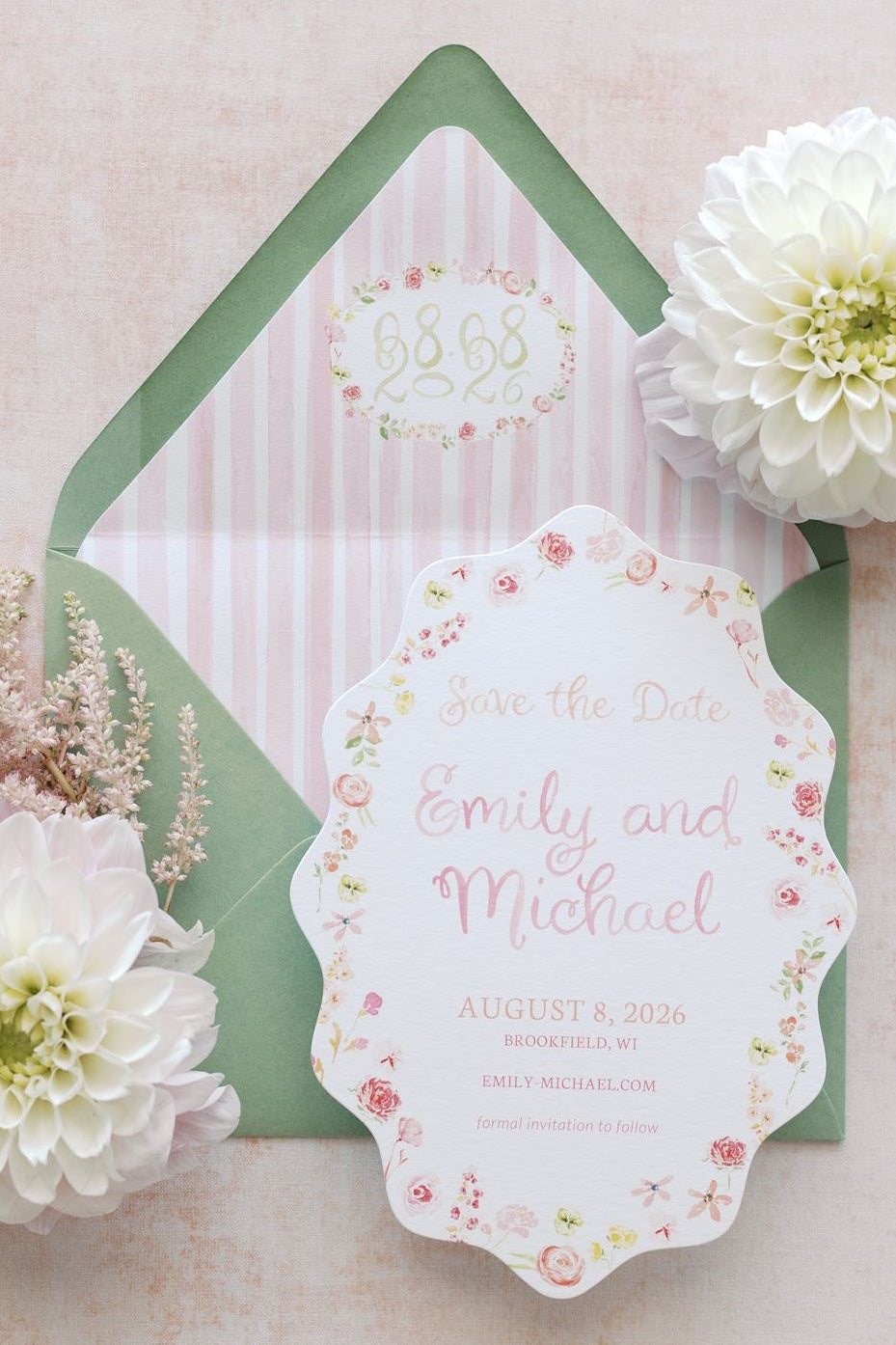 A styled flat-lay of a wedding invitation card with a scalloped floral border, placed in front of a green and pink striped envelope. The arrangement is decorated with two large white dahlia flowers and delicate pink filler stems on a soft blush pink textured background.