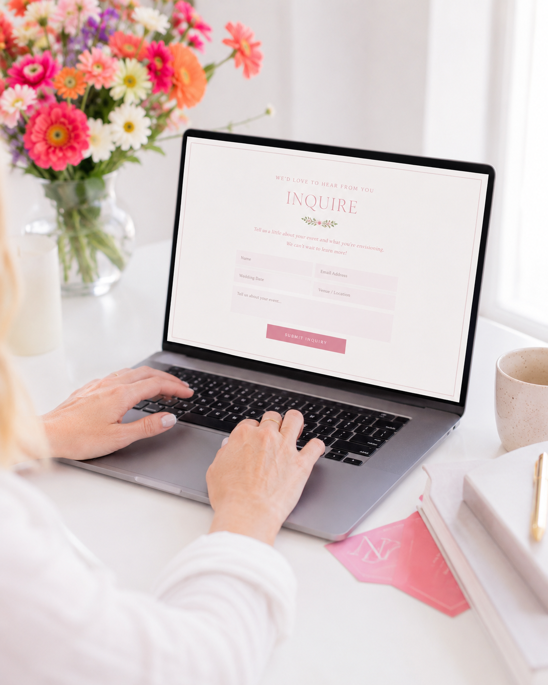 A person typing on a laptop at a bright desk, with a website inquiry form visible on the screen. A vase of colorful flowers, a mug, and stacked books are on the desk.