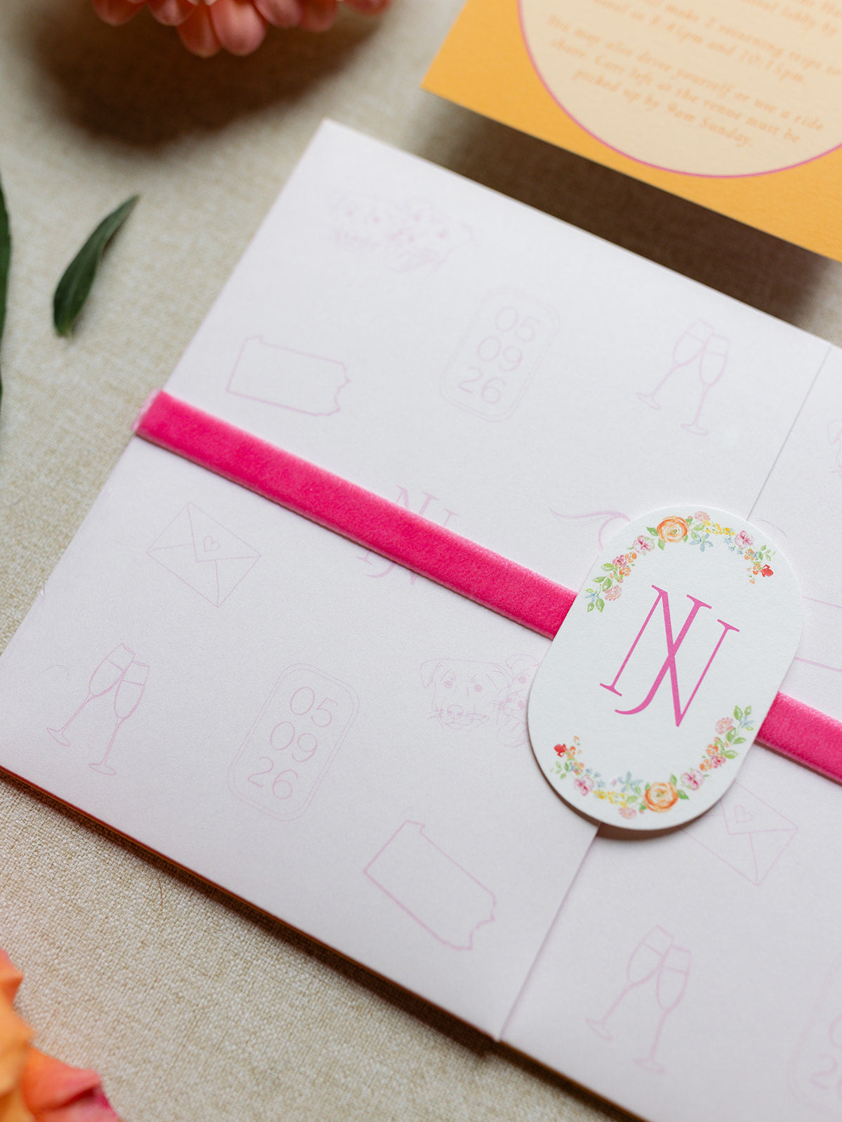 A close-up of a white invitation card or envelope wrapped with a bright pink velvet ribbon and a round white sticker seal featuring a floral wreath and the monogram 'NJ'.