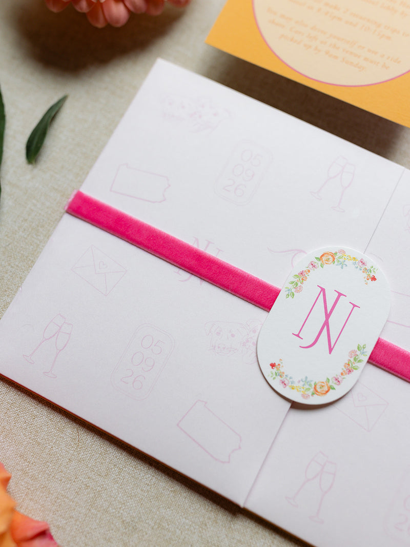 A close-up of a white invitation card or envelope wrapped with a bright pink velvet ribbon and a round white sticker seal featuring a floral wreath and the monogram 'NJ'.